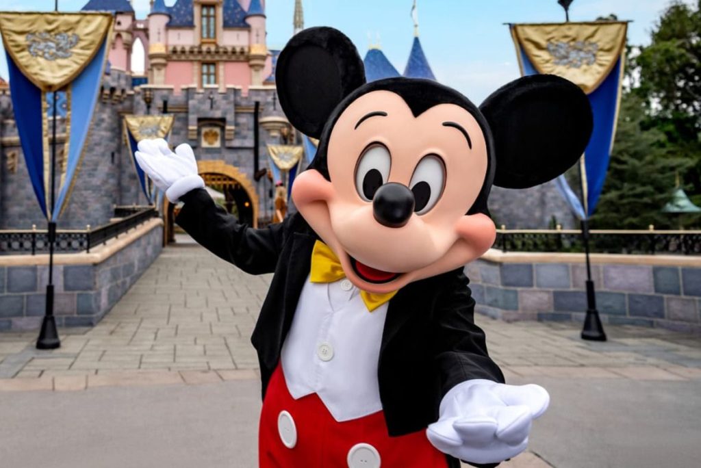 A cheerful character in a tuxedo and bow tie poses in front of a fairytale castle, welcoming visitors with open arms in a vibrant park setting, featured in the Disney Vacation Club Stay Your Way Sweepstakes