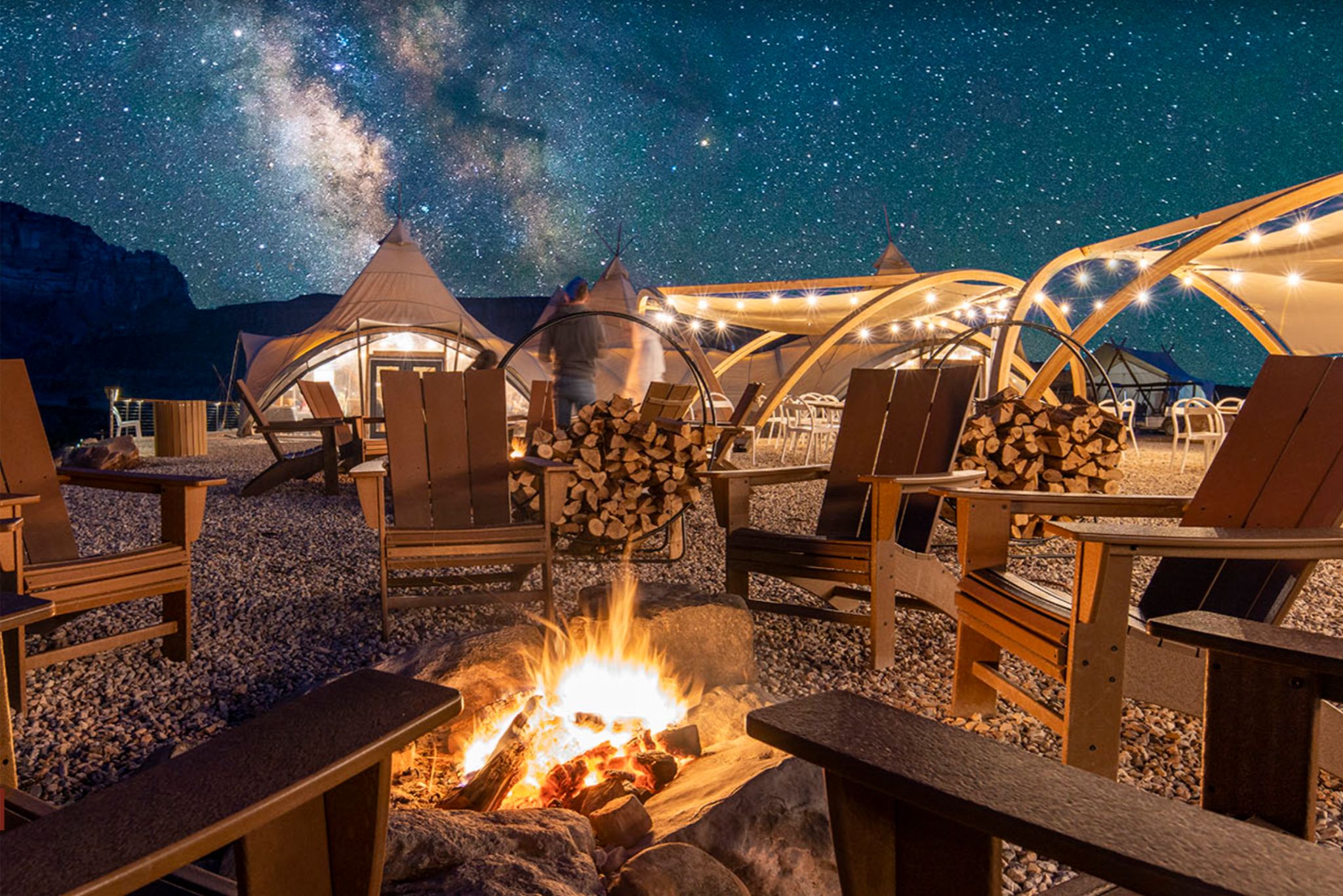A cozy outdoor seating area features wooden chairs surrounding a glowing campfire, with illuminated tents and a starry sky providing a serene backdrop, featured in the Utah Under Canvas Sweepstakes