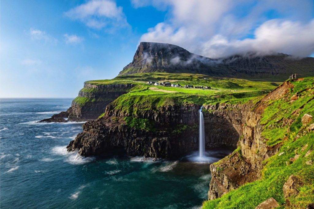 A lush, green cliffside overlooks a serene ocean, where a waterfall cascades into the water below. Clouds drift over a distant mountain, enhancing the tranquil landscape, featured in the Icelandair Bad Photographer Sweepstakes