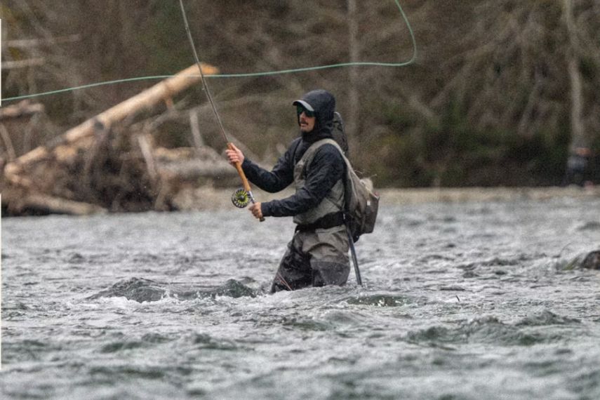 A fisherman stands in a river, casting a fly rod while wearing waders. Trees and logs are visible in the background, suggesting a natural fishing environment, featured in the Sportsmans Warehouse Fishing Trip Sweepstakes