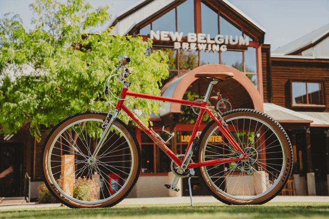 A bright red bicycle stands in front of a rustic brewery with large glass windows, surrounded by green trees and grass under a clear blue sky, featured in the 2026 National Bike Giveaway