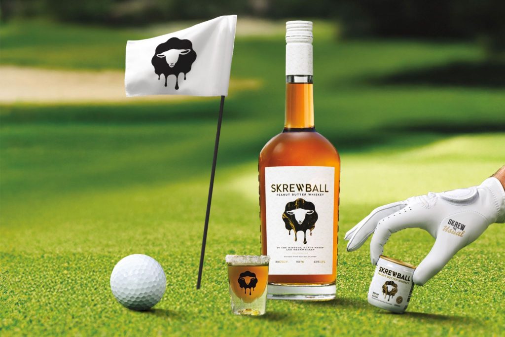 A golf ball rests on green grass next to a bottle and shot glass of whiskey, while a hand in a golf glove reaches for a small can nearby. A flag with a sheep design stands in the background, featured in the Skrewball $25,000 Sweepstakes