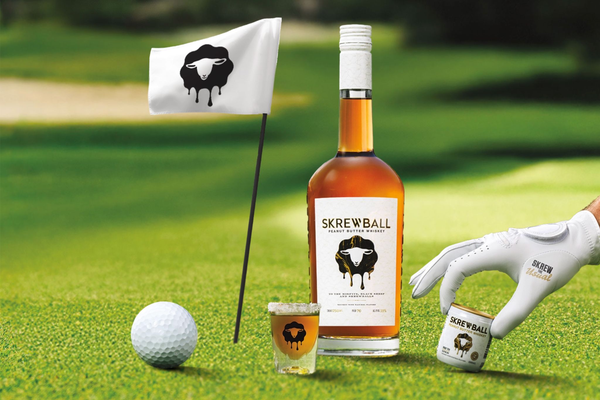 A golf ball rests on green grass next to a bottle and shot glass of whiskey, while a hand in a golf glove reaches for a small can nearby. A flag with a sheep design stands in the background, featured in the Skrewball $25,000 Sweepstakes