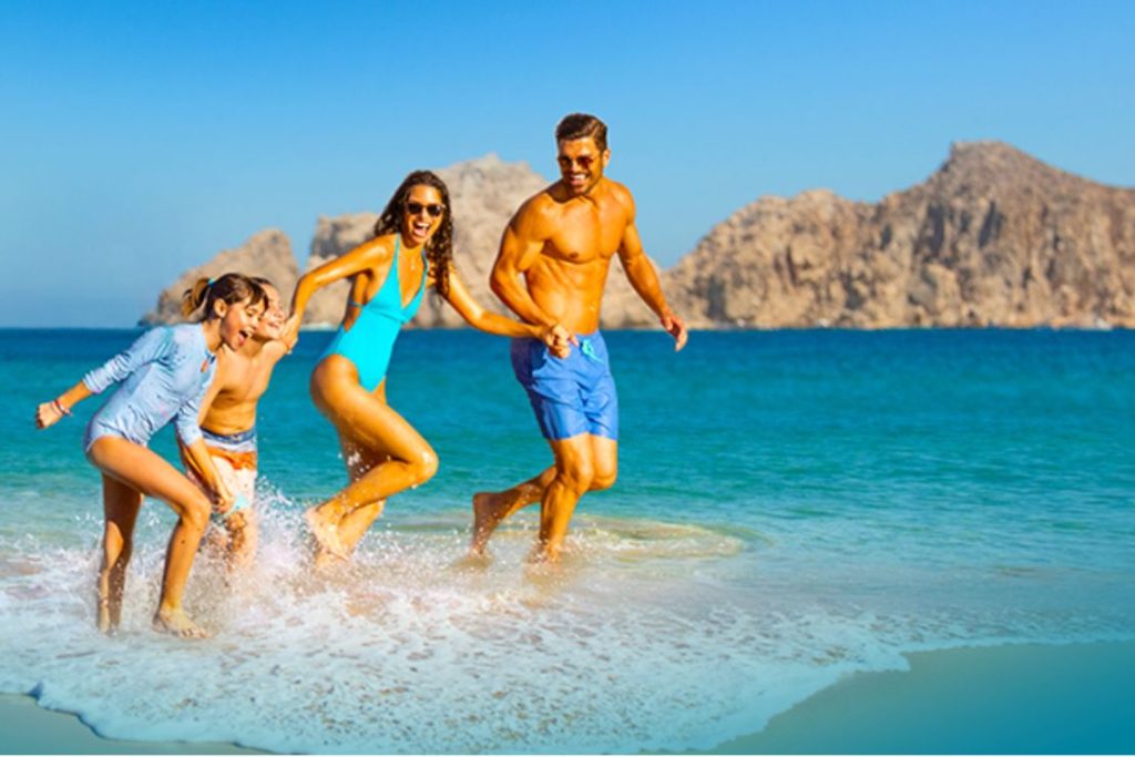A family joyfully splashes through shallow water at a beach, with a sunny sky and rocky formations in the background, capturing a moment of summer fun and togetherness, featured in the BJ's Travel Grand Giveaway
