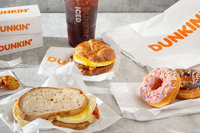 A spread of breakfast items includes a croissant sandwich with eggs and cheese, a toasted bread sandwich with eggs, and colorful donuts alongside a cold iced beverage, all on a light surface, featured in the Dunkin' Cart Catch Sweepstakes