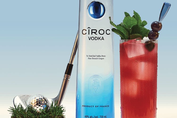 A stylish vodka bottle stands beside a cocktail garnished with mint and berries, with a golf club and ball resting on green grass, creating a refreshing summer vibe, featured in the CIROC TGL Golf Finals Trip Sweepstakes