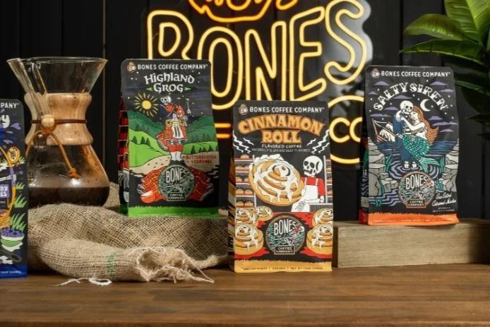 A wooden table displays several colorful coffee bags with unique illustrations, alongside a glass coffee maker and burlap, set against a dark background with neon lighting, featured in the Bones Coffee Free Coffee Sweepstakes