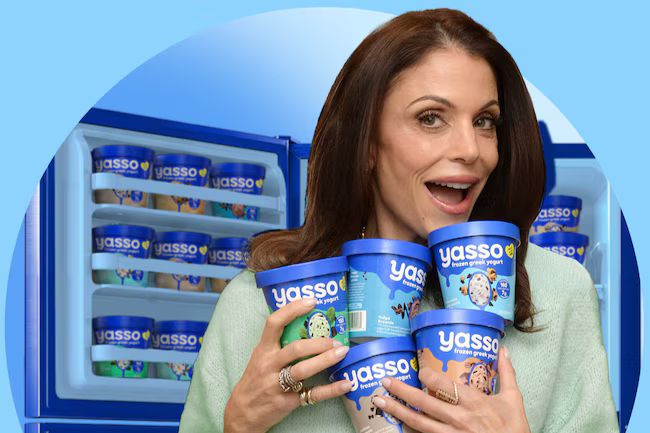 A woman smiles while holding multiple containers of frozen yogurt, surrounded by a freezer filled with more of the same brand. Bright blue backdrop enhances the scene, featured in the Yasso Foolproof Freezer Giveaway