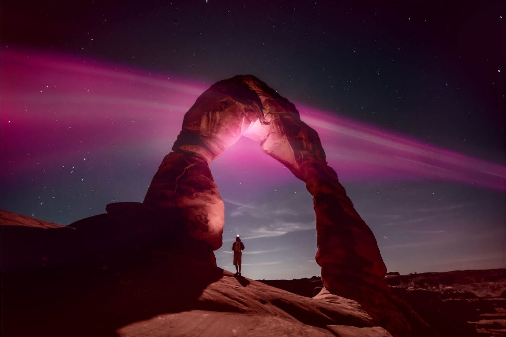A lone figure stands beneath a natural rock arch at night, illuminated by pink light against a starry sky, creating a striking contrast with the surrounding landscape, featured in the T-Mobile National Park Trip Sweepstakes
