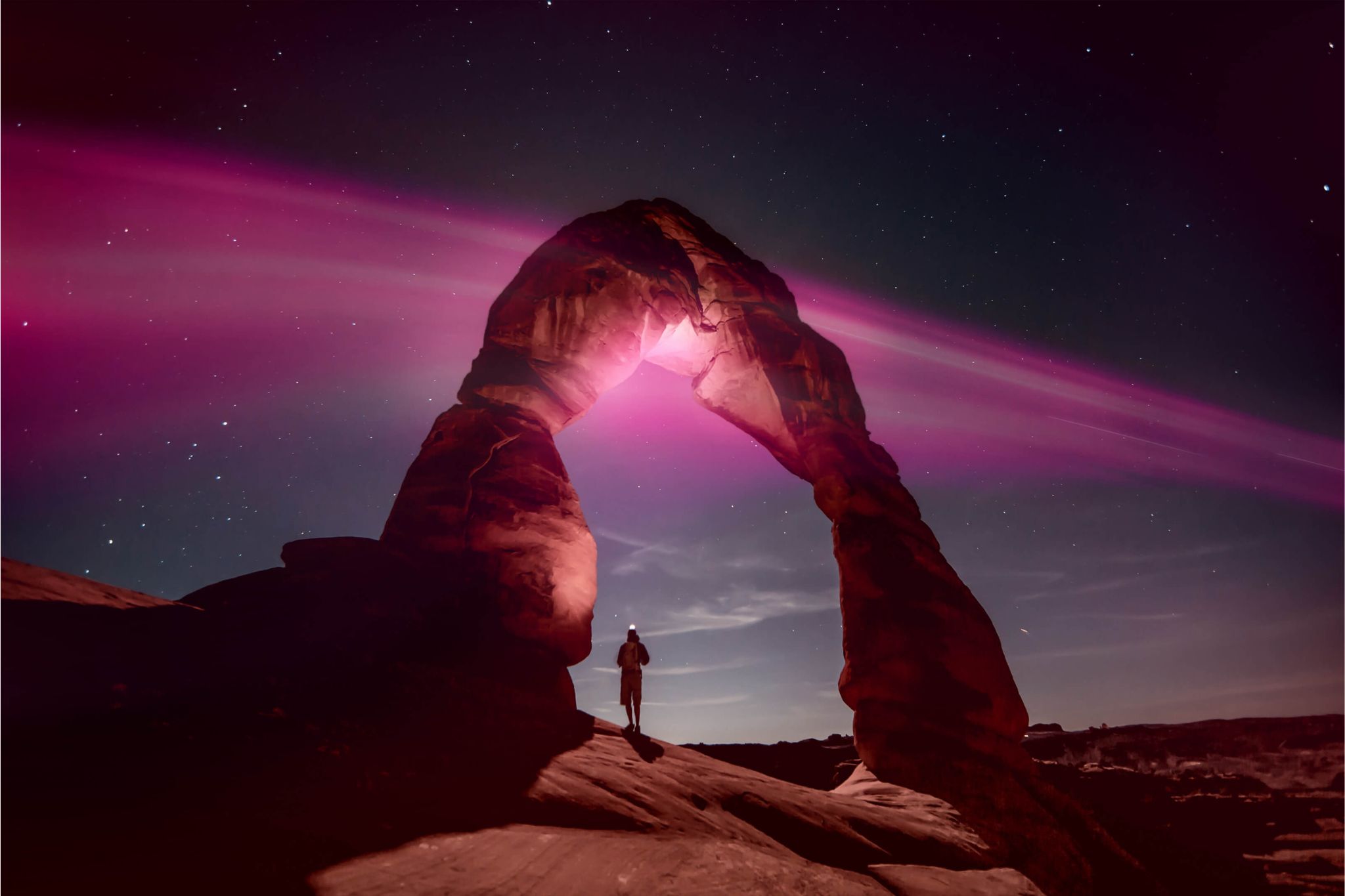 A lone figure stands beneath a natural rock arch at night, illuminated by pink light against a starry sky, creating a striking contrast with the surrounding landscape, featured in the T-Mobile National Park Trip Sweepstakes