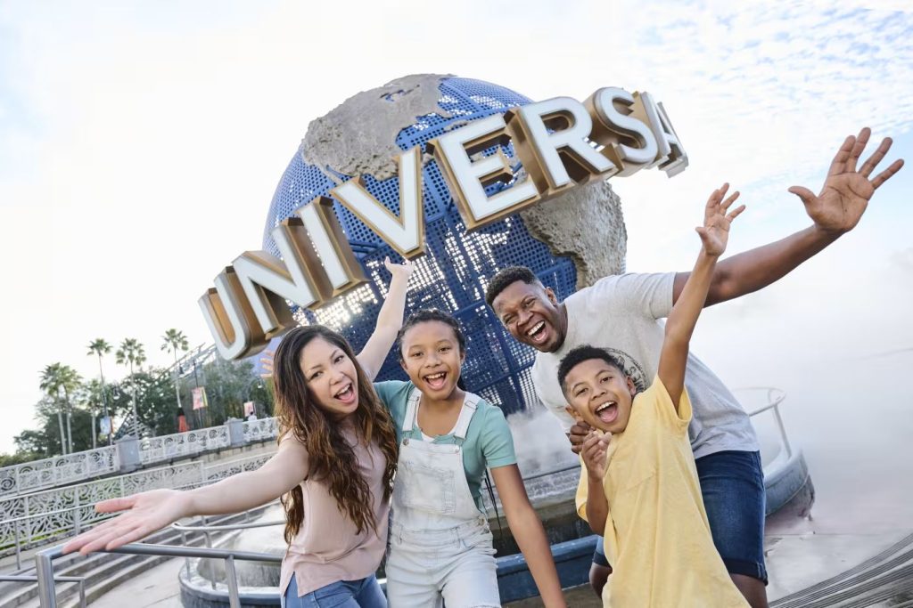 A joyful family poses in front of a large globe sculpture, celebrating at a theme park. They are smiling and making fun gestures, capturing a moment of excitement and happiness, featured in the Visit Orlando Family Vacation Sweepstakes