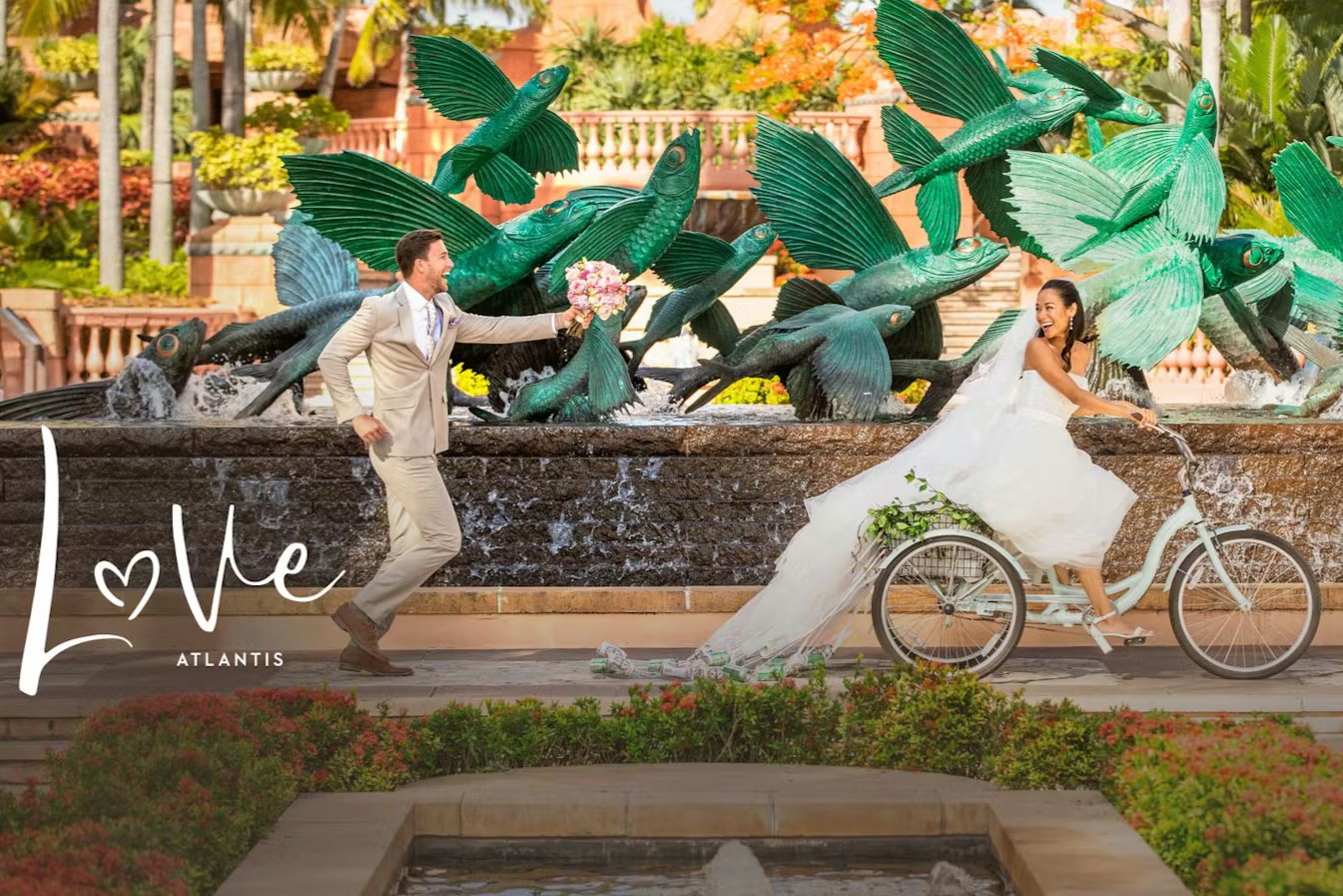 A bride happily rides a bicycle near a vibrant fountain adorned with fish sculptures, while the groom playfully reaches out with a bouquet, set in a lush, romantic garden, featured in the Atlantis Bahamas Wedding Sweepstakes