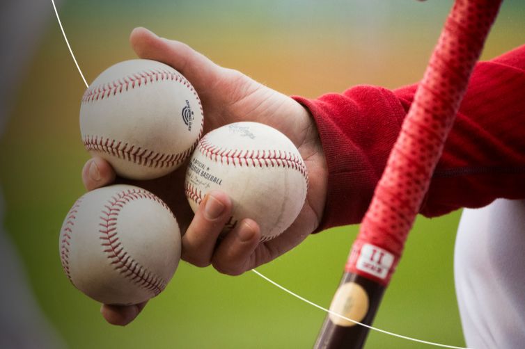 A hand holds three baseballs, preparing for a pitch, with a bat resting nearby; the scene captures the anticipation of a baseball game, featured in the Mastercard MLB Postseason Sweepstakes