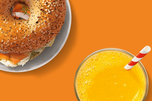 A sesame seed bagel topped with smoked salmon and cream cheese sits on a white plate next to a glass of orange juice with a striped straw, all against a vibrant orange background, featured in the Tropicana Spotify Premium Sweepstakes