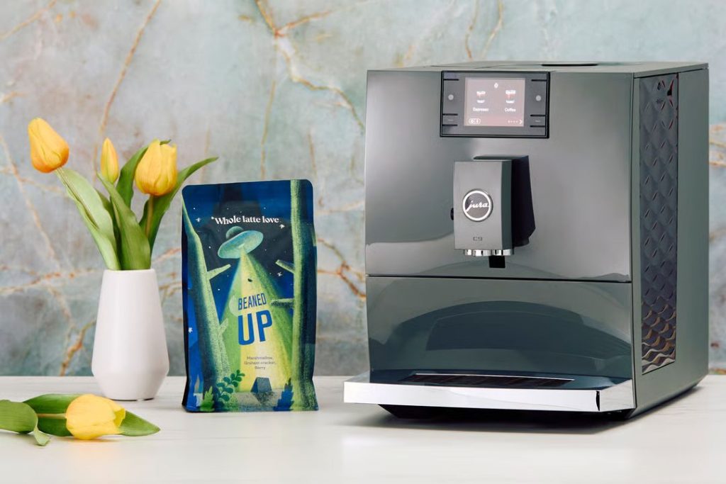 A sleek coffee machine sits on a countertop beside a colorful bag of coffee. Fresh yellow tulips in a white vase add a pop of color to the modern, stylish setting, featured in the Whole Latte Love JURA C9 Sweepstakes