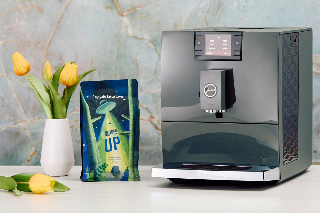 A sleek coffee machine sits on a countertop beside a colorful bag of coffee. Fresh yellow tulips in a white vase add a pop of color to the modern, stylish setting, featured in the Whole Latte Love JURA C9 Sweepstakes