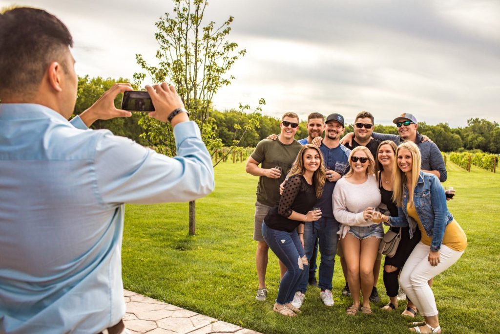 A group of nine friends poses joyfully for a photo in a lush, green vineyard, while a person in a light blue shirt captures the moment with a smartphone, featured in the Visit Beloit Weekend Getaway Sweepstakes