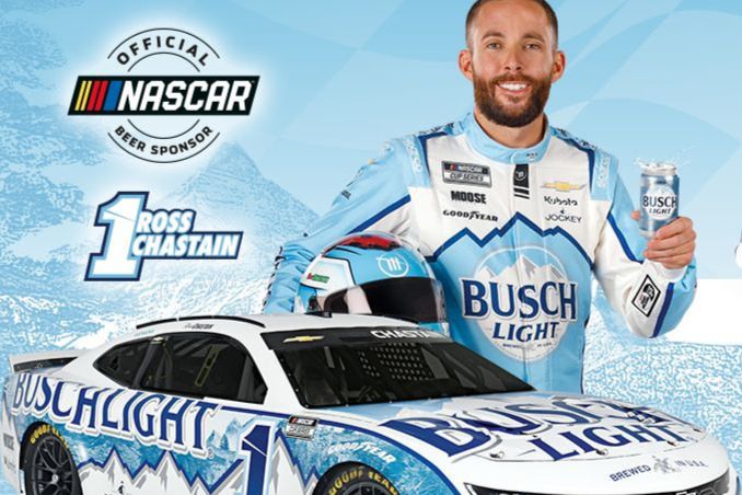 A smiling man in a blue racing suit holds a can while standing beside a white race car decorated in blue and white. The background features a light blue design, evoking a festive atmosphere, featured in the Busch Racing Sweepstakes