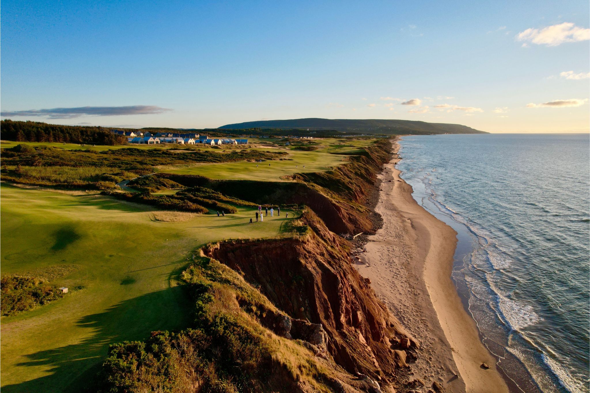 A scenic coastal golf course with lush green fairways and steep cliffs overlooking the shoreline, where golfers are playing near the edge, with a calming sunset in the background, featured in the Cabot Golf Trip Sweepstakes