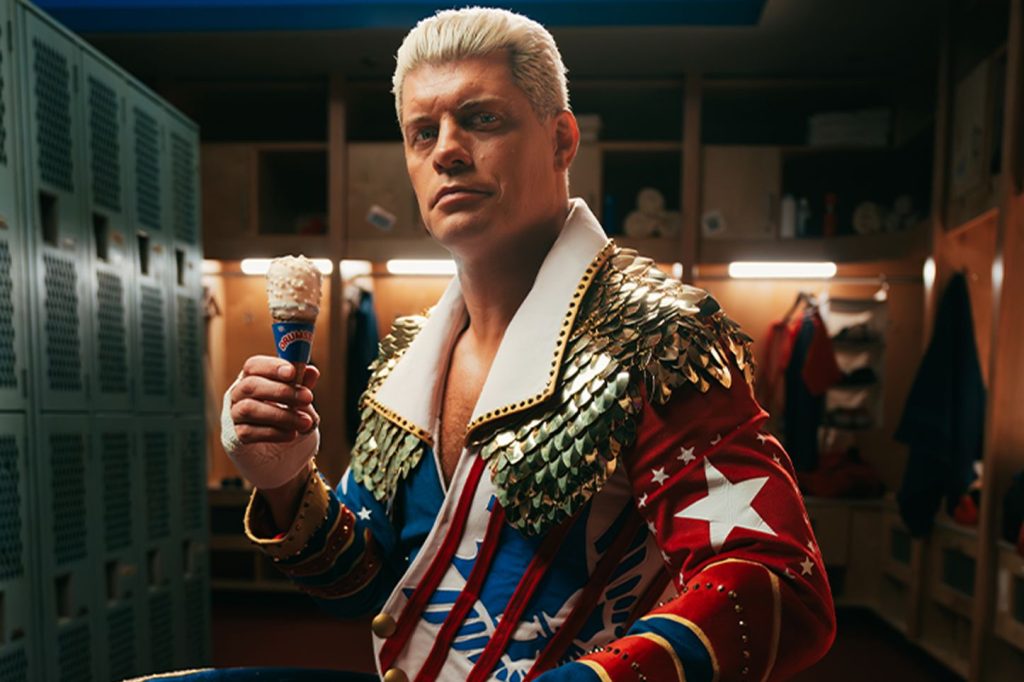 A confident man in a colorful, ornate outfit holds an ice cream cone while standing in a locker room, showcasing a blend of athleticism and playfulness, featured in the Drumstick Summer Snackdown Sweepstakes