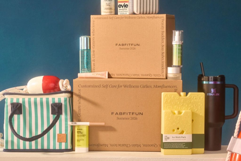 A colorful assortment of beauty and wellness products is displayed on stacked boxes, alongside a striped cooler bag and a travel mug, creating an inviting self-care arrangement, featured in the FabFitFun Summer Dream Sweepstakes