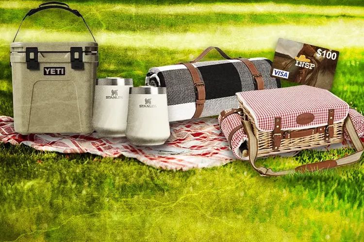 A cozy outdoor setup features a cooler, two drink tumblers, a picnic blanket, a wicker basket, and a gift card, all arranged on a grassy area, suggesting a perfect picnic scene, featured in the INSP Prairie Picnic Sweepstakes