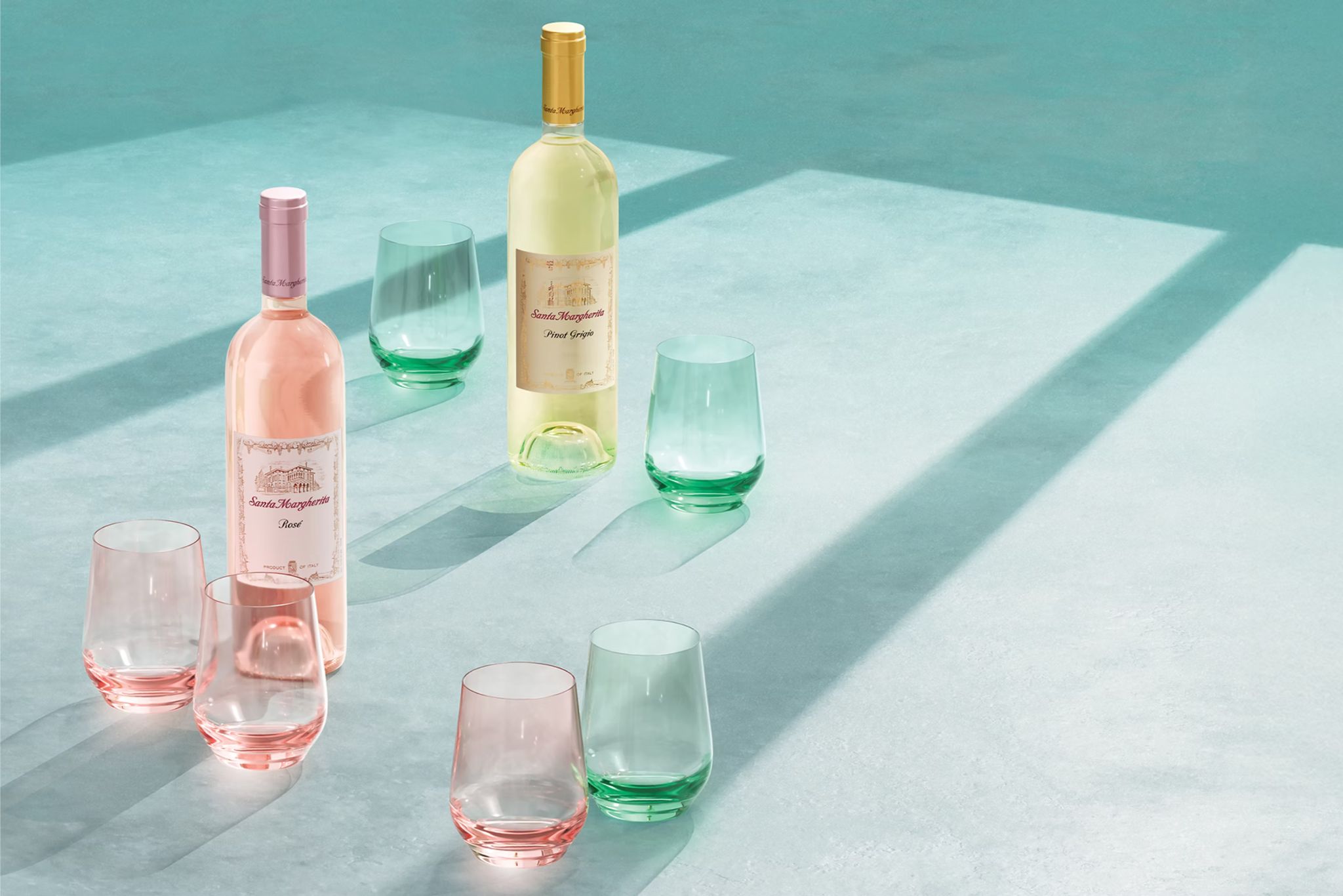Two bottles of wine, one pink and one white, sit among colorful glassware on a pale blue surface, with soft shadows creating a serene, inviting atmosphere, featured in the Santa Margherita Estelle Glass Sweepstakes