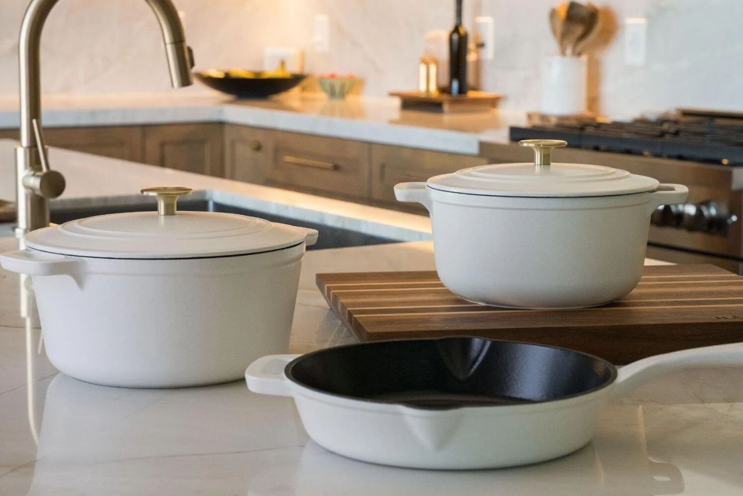 A modern kitchen countertop features two white cooking pots with lids and one black skillet, arranged neatly on a wooden cutting board with a warm, inviting atmosphere, featured in the Mother’s Day Cast Iron Giveaway