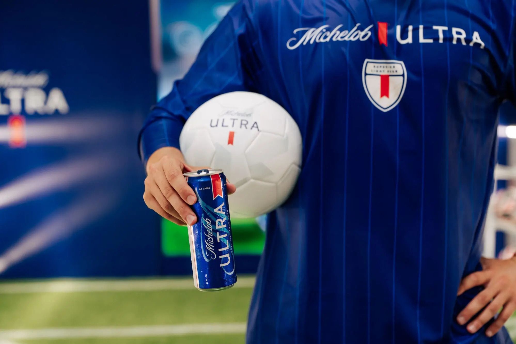 A person in a blue jersey holds a soccer ball under one arm while gripping a can of beer in the other hand, standing on a green turf field with an out-of-focus background, featured in the Michelob Ultra Chief Trophy Officer Sweepstakes