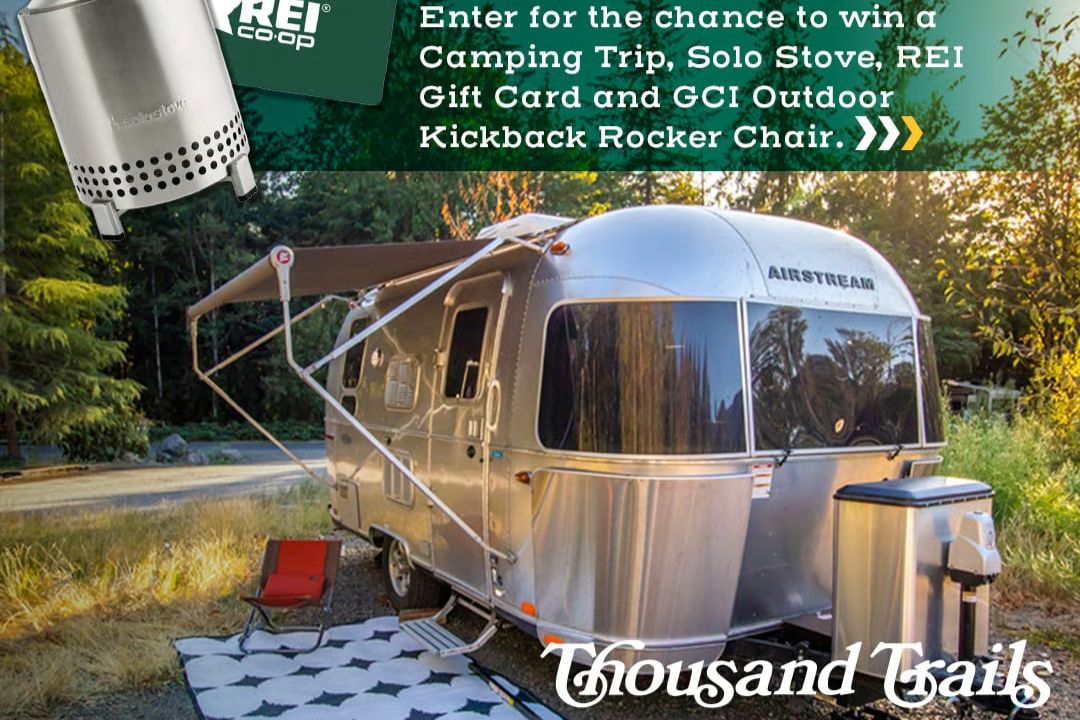A shiny Airstream trailer is parked in a wooded area, with a cozy outdoor seating setup, inviting a relaxing camping experience surrounded by nature, featured in the Gear Up and Camp Sweepstakes
