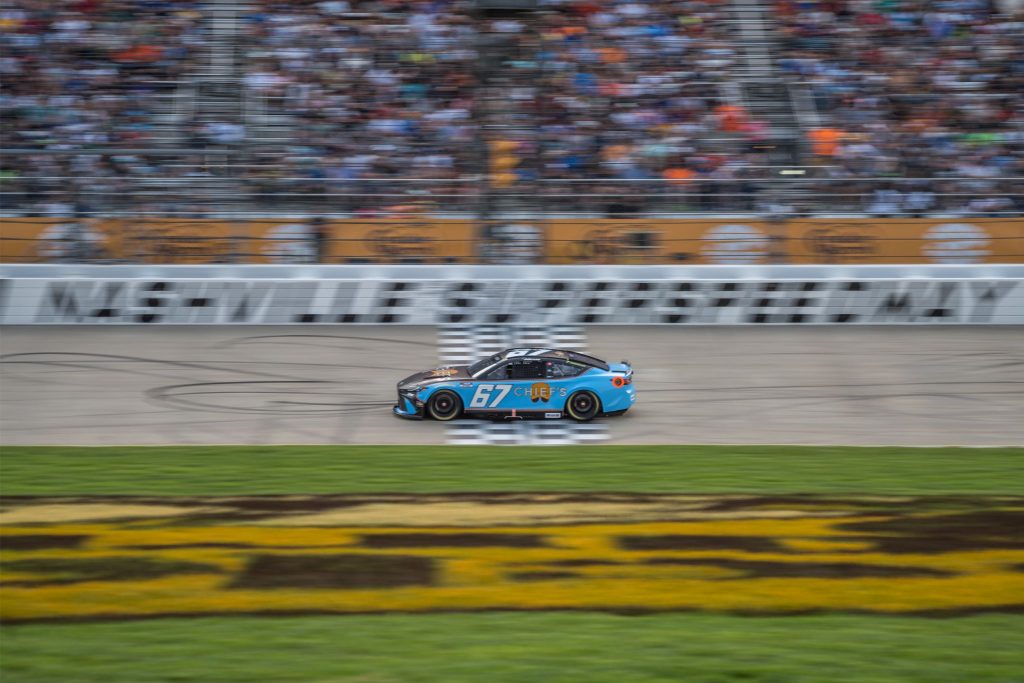 A blue and black race car speeds around a racetrack, leaving tire marks on the asphalt, with a cheering crowd visible in the blurred background, featured in the Field & Stream Nashville Race Weekend Sweepstakes