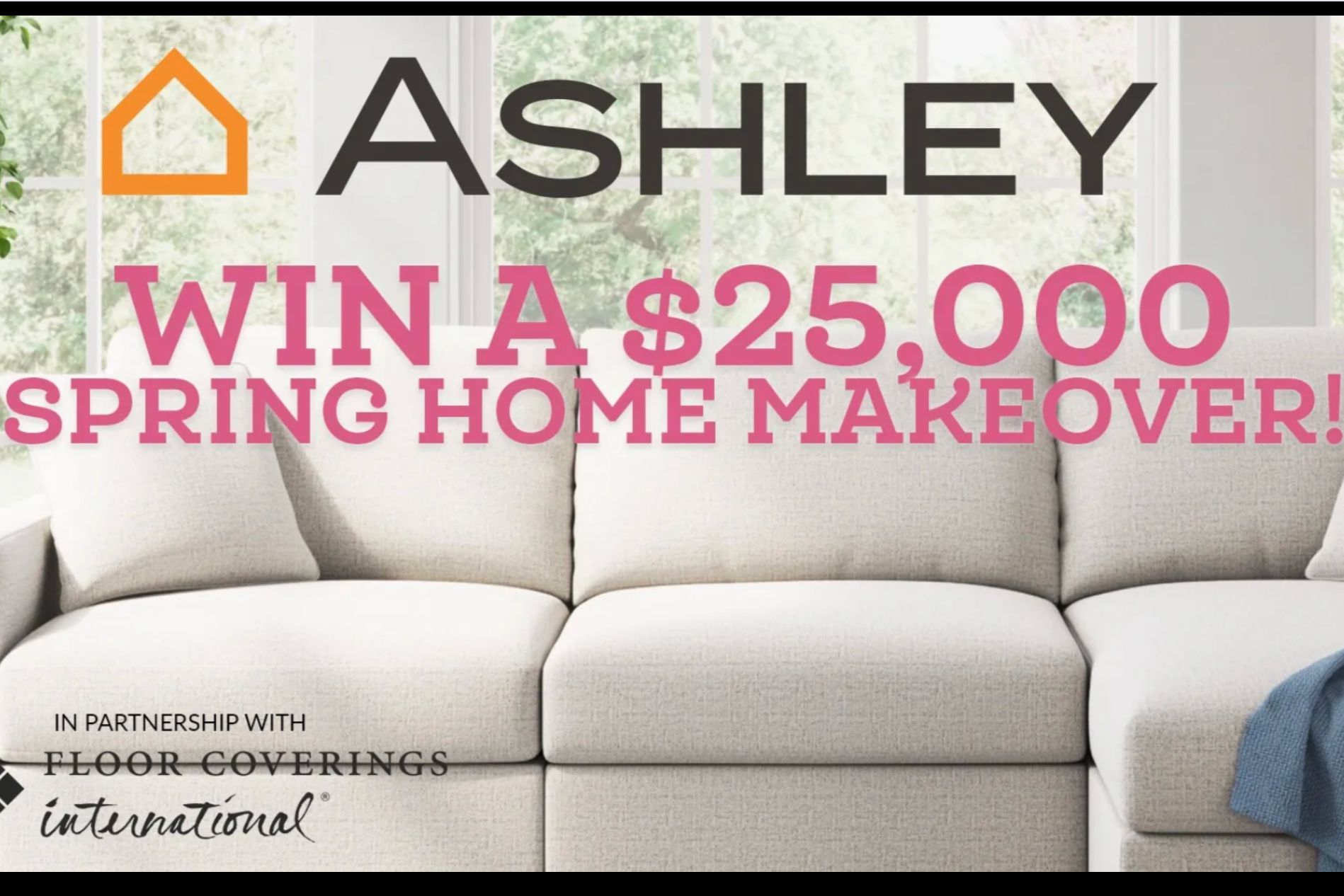 A cozy light-colored sofa with plush cushions is featured prominently, set against a backdrop of large windows letting in natural light, creating a warm and inviting atmosphere, featured in the Ashley Home Makeover Sweepstakes