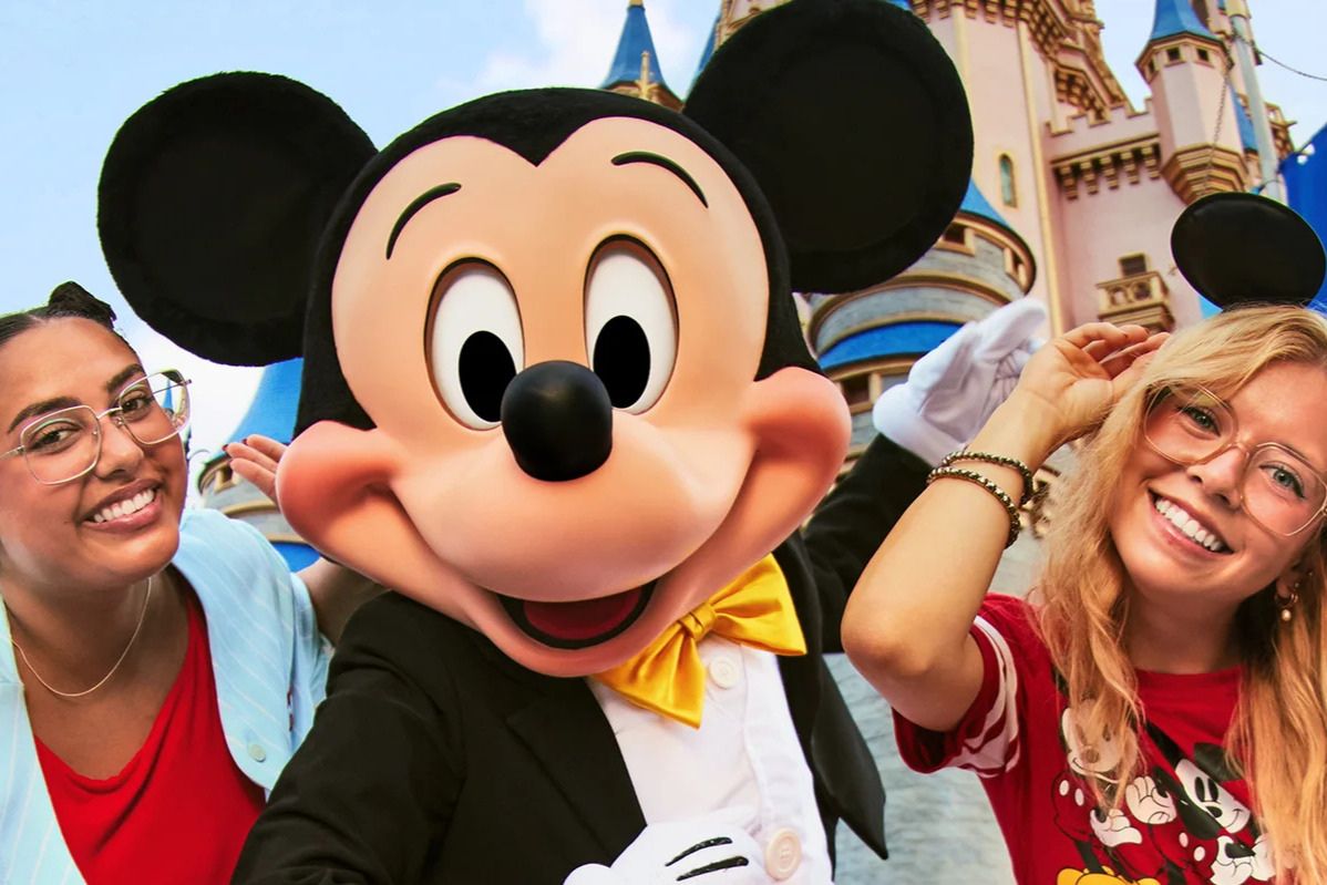 Two smiling women pose with a cheerful costumed character at a theme park, surrounded by a colorful castle backdrop, enjoying a fun moment together, featured in the Upgraded Points Ultimate Summer of Magic Sweepstakes