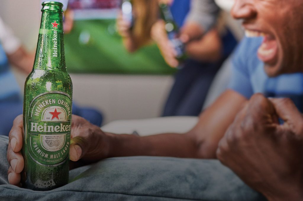 A person joyfully holds a green beer bottle while cheering with friends in a lively setting, celebrating alongside a blurred background that suggests a sports event, featured in the Heineken Kansas City Soccer Sweepstakes