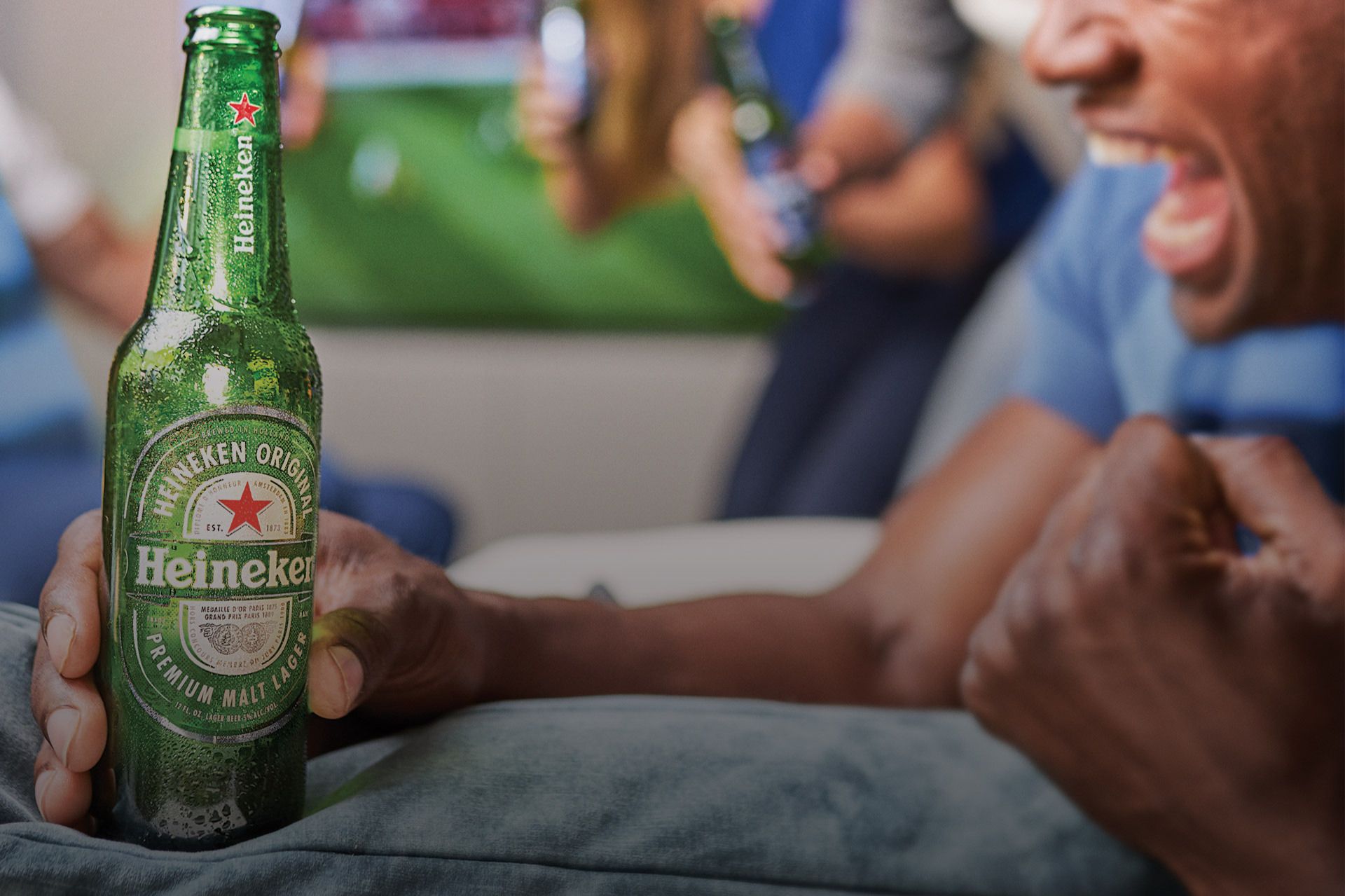 A person joyfully holds a green beer bottle while cheering with friends in a lively setting, celebrating alongside a blurred background that suggests a sports event, featured in the Heineken Kansas City Soccer Sweepstakes