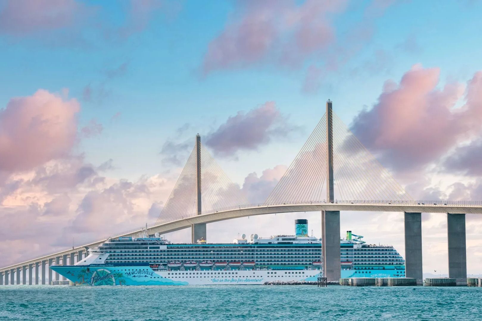 A large cruise ship passes under a modern cable-stayed bridge, surrounded by calm waters and a pastel sky filled with soft clouds, featured in the Margaritaville at Sea 7-Night Cruise Sweepstakes