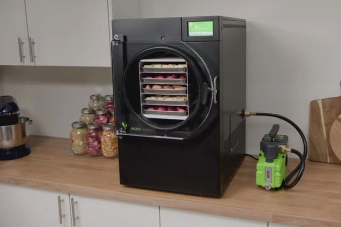 A black freeze dryer sits on a wooden countertop, showcasing food trays inside, with colorful jars of preserved snacks in the background and a small pump connected, featured in the Harvest Right Freeze Dryer Sweepstakes