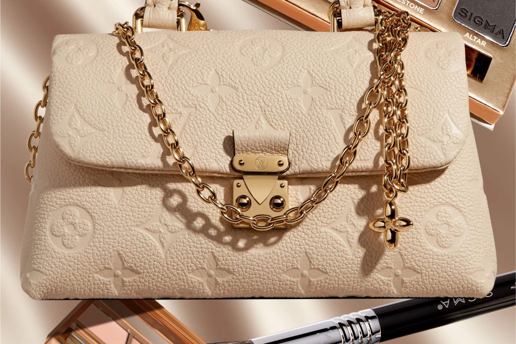 A stylish beige handbag with a gold chain strap and clasp showcases intricate embossed patterns, set against a soft backdrop with beauty products partially visible, featured in the Sigma Beauty Louis Vuitton Nano Madeleine Sweepstakes