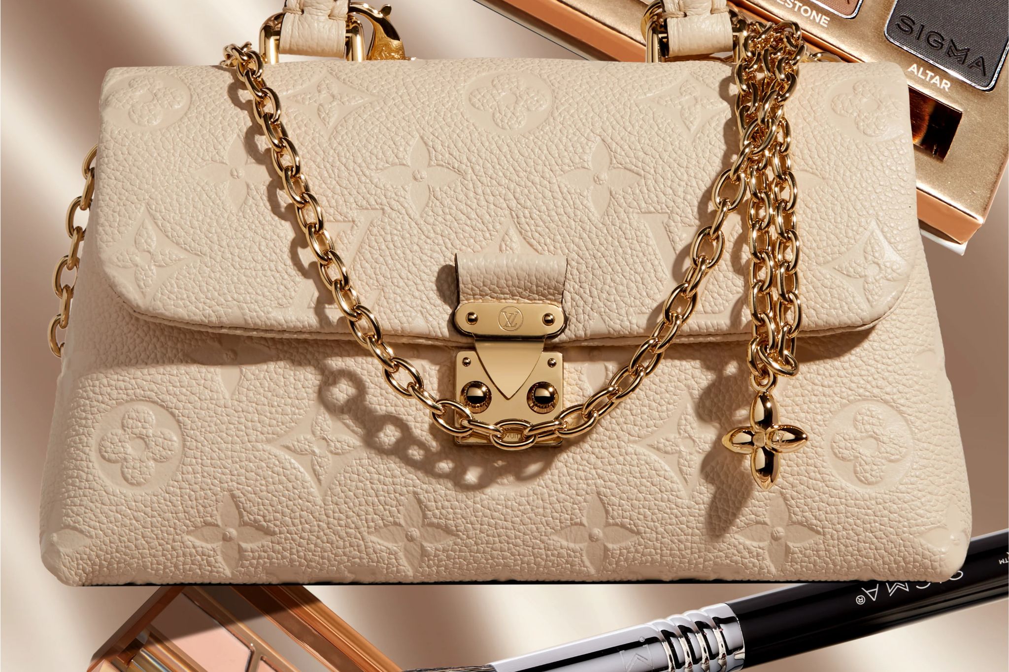 A stylish beige handbag with a gold chain strap and clasp showcases intricate embossed patterns, set against a soft backdrop with beauty products partially visible, featured in the Sigma Beauty Louis Vuitton Nano Madeleine Sweepstakes