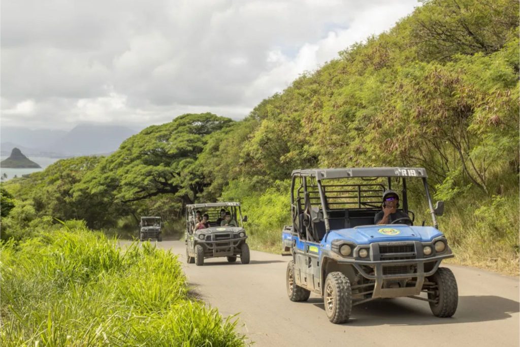 A convoy of all-terrain vehicles drives along a dirt road surrounded by lush greenery, with a scenic view of hills and a body of water in the background, featured in the Hawaii.com Oahu Family Vacation Sweepstakes