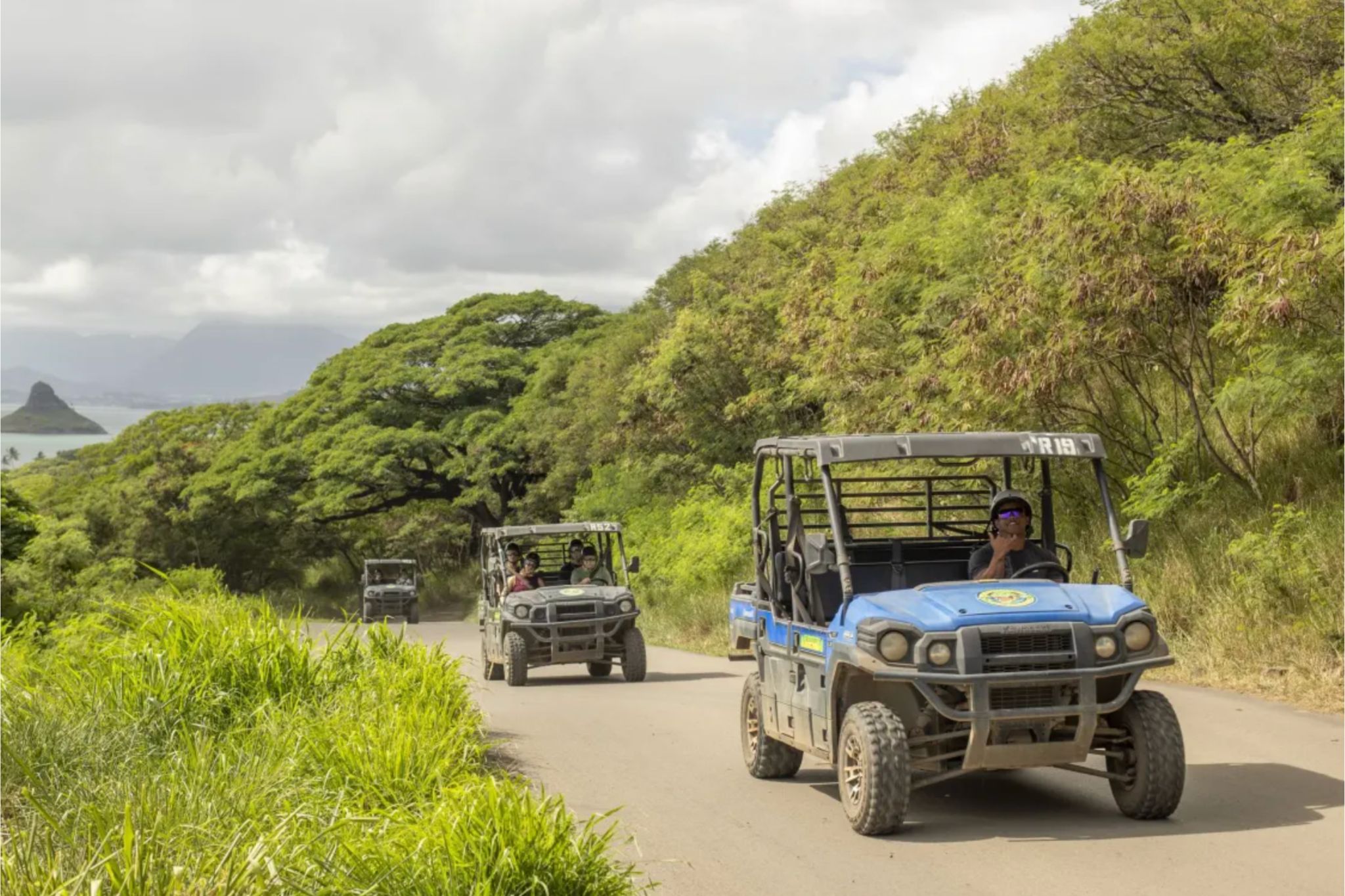 A convoy of all-terrain vehicles drives along a dirt road surrounded by lush greenery, with a scenic view of hills and a body of water in the background, featured in the Hawaii.com Oahu Family Vacation Sweepstakes