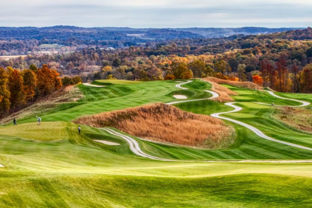 Rolling green hills of a golf course surrounded by autumn trees, with a few golfers in the distance enjoying a game under a cloudy sky, featured in the French Lick Resort Luxury Getaway
