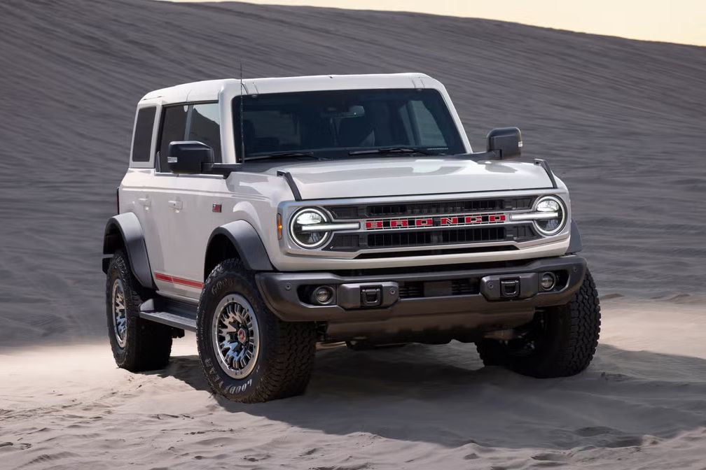 A rugged SUV is parked on a sandy desert landscape, showcasing its bold design and off-road capabilities. The vehicle features prominent headlights and large, textured tires, featured in the Ford Bronco Badlands Sweepstakes