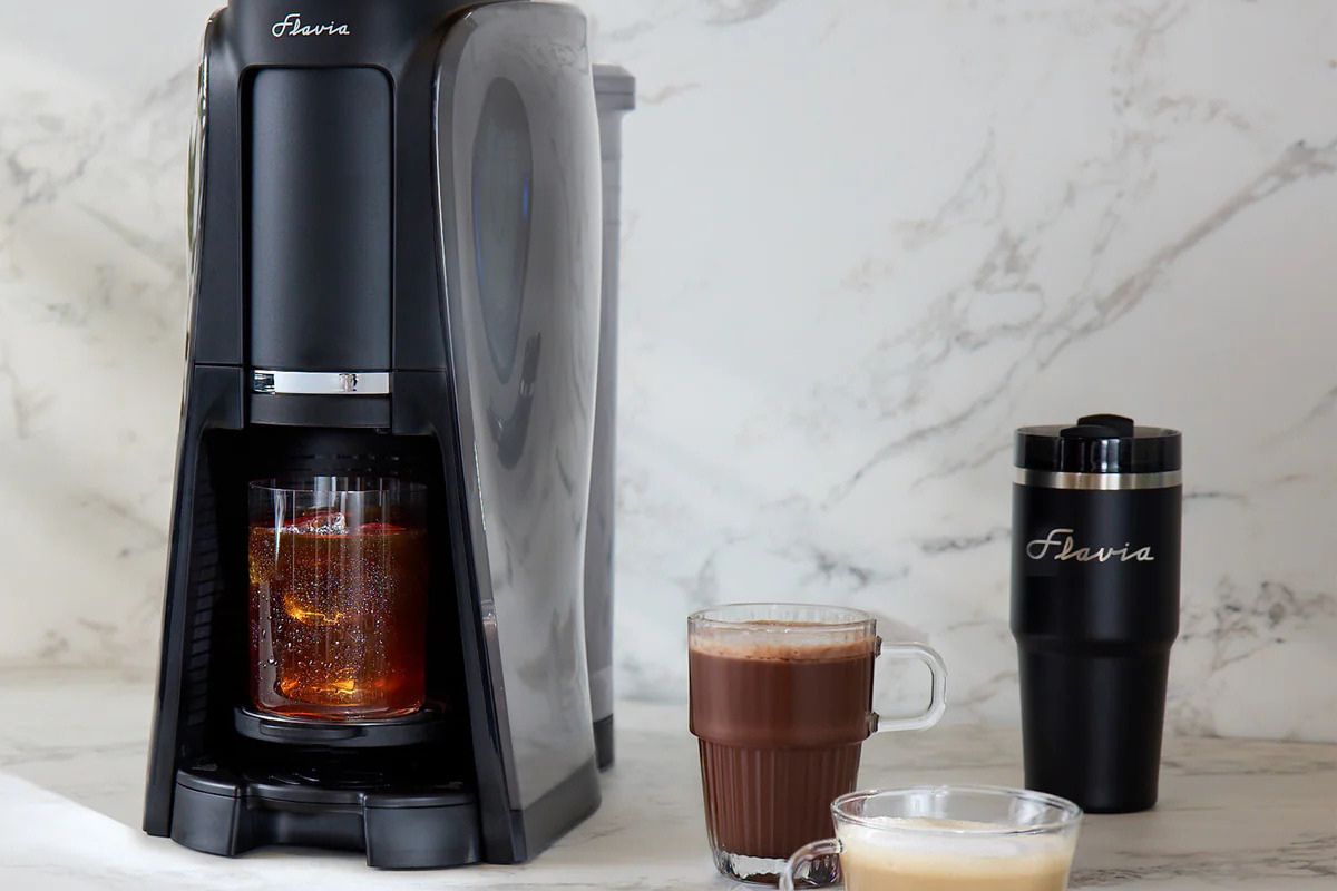 A sleek coffee maker brews a cup while two mugs and a travel tumbler sit nearby, showcasing a rich beverage juxtaposed against a light marble background, featured in the Flavia AROMA Bundle Sweepstakes