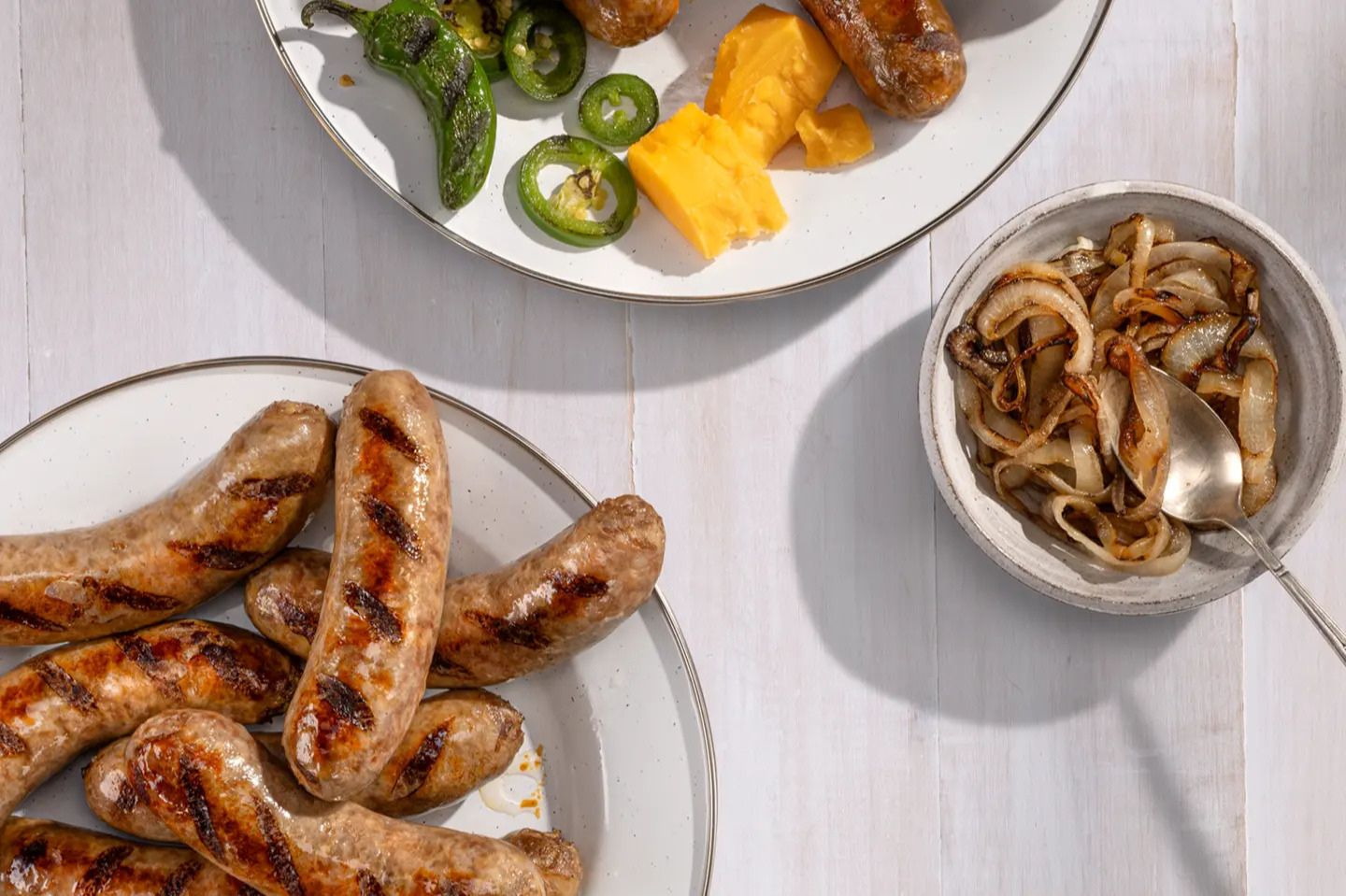 Grilled sausages rest on a white plate, accompanied by jalapeños, cheese, and grilled onions in a bowl nearby, creating a delicious, inviting spread, featured in the Smithfield Backyard Bratluck Sweepstakes