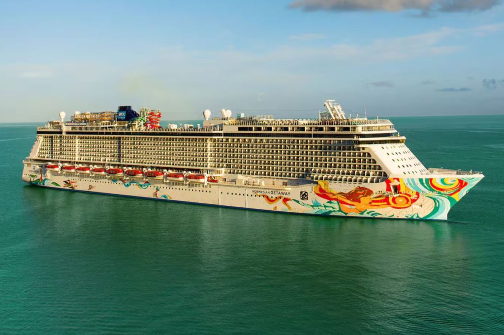 A large cruise ship with a colorful abstract design sails smoothly on calm blue waters under a clear sky, showcasing levels of decks and various attractions, featured in the Elvis Duran Cruise Sweepstakes
