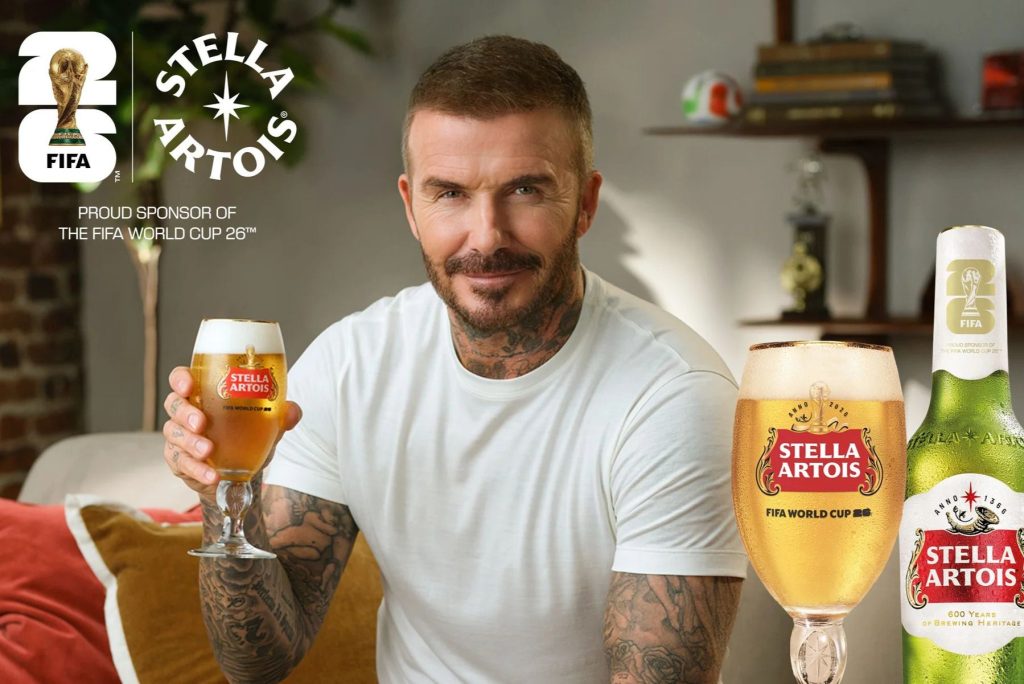 A man with tattoos smiles while holding a glass of beer, seated on a couch with a cozy atmosphere. Two beer bottles are visible next to him, featured in the Stella Artois FIFA World Cup 2026 Sweepstakes