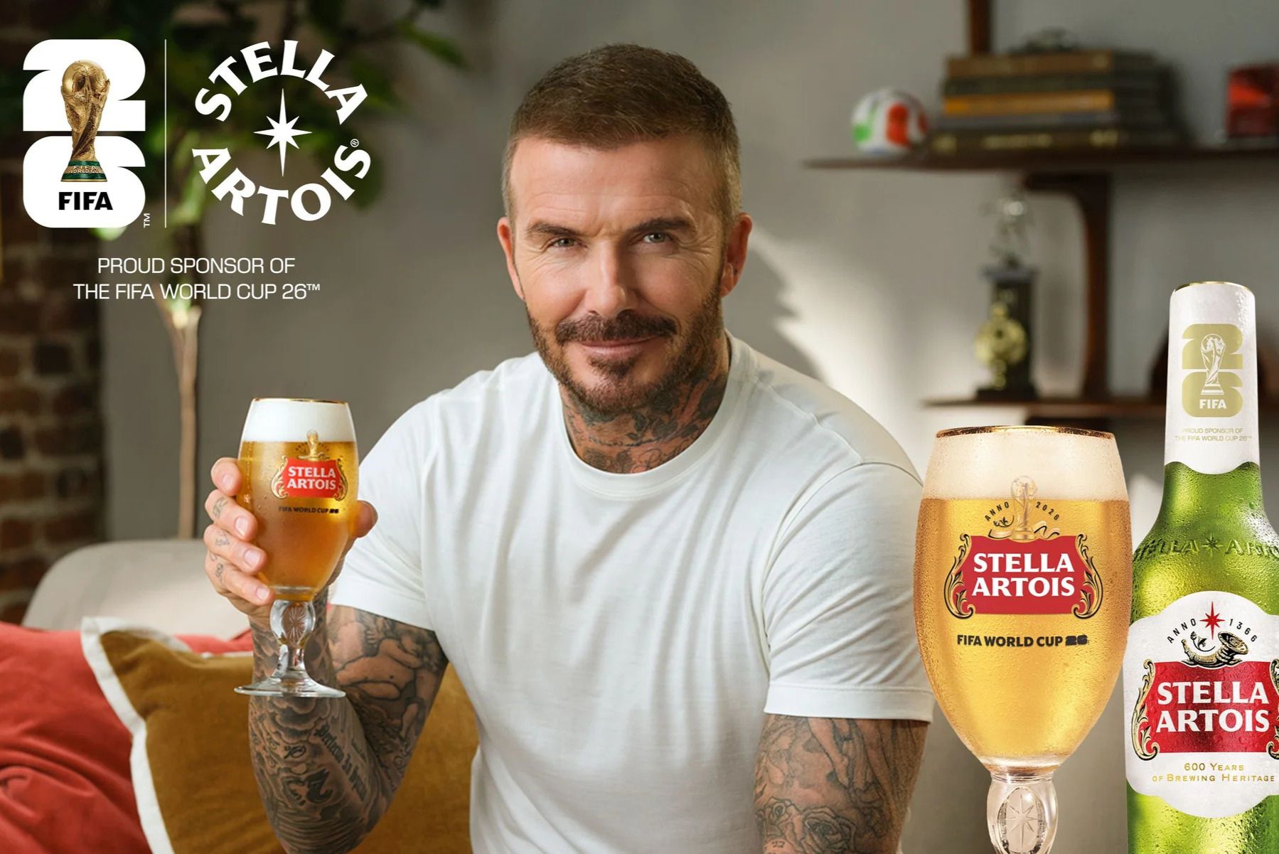 A man with tattoos smiles while holding a glass of beer, seated on a couch with a cozy atmosphere. Two beer bottles are visible next to him, featured in the Stella Artois FIFA World Cup 2026 Sweepstakes