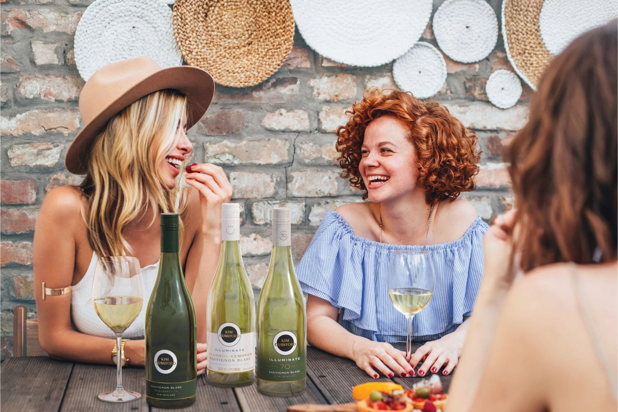 Two friends share laughter at a wooden table, enjoying wine and snacks, surrounded by a rustic brick wall and decorative woven plates. One woman playfully eats a berry, featured in the Kim Crawford New Zealand Trip Sweepstakes