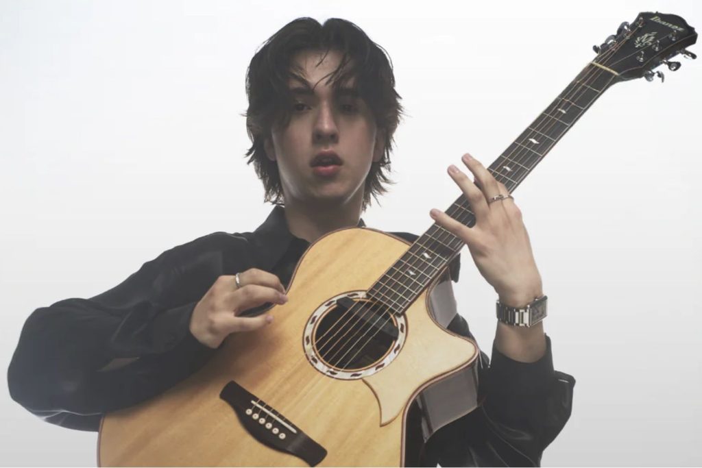 A young man with tousled hair plays an acoustic guitar, dressed in a black shirt. He gazes directly at the camera, holding the guitar with focused intent, featured in the Daddario Marcin Guitar Giveaway